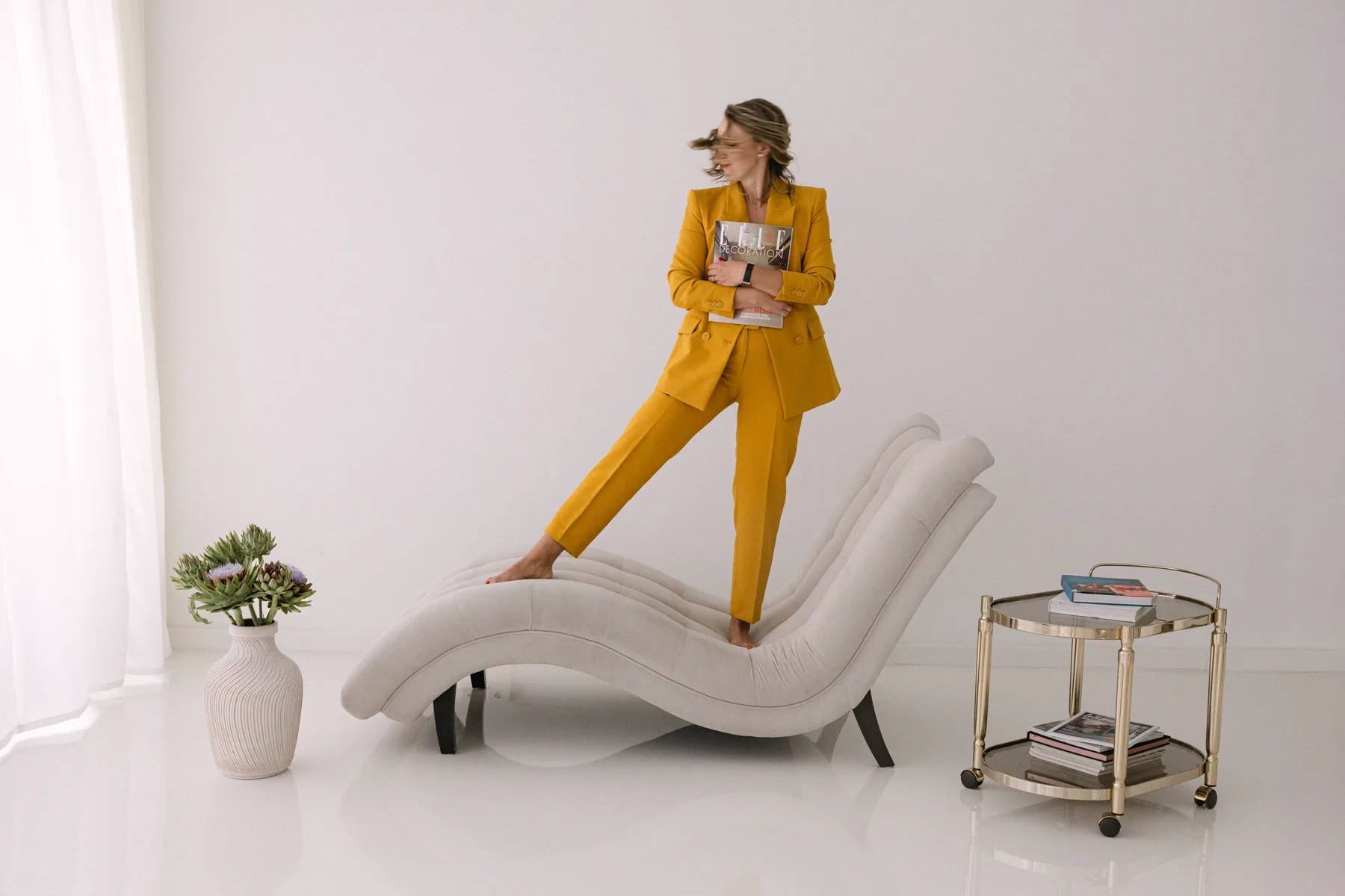 Adrienn Beck in a yellow suit beside a white chaise lounge with an interior magazine